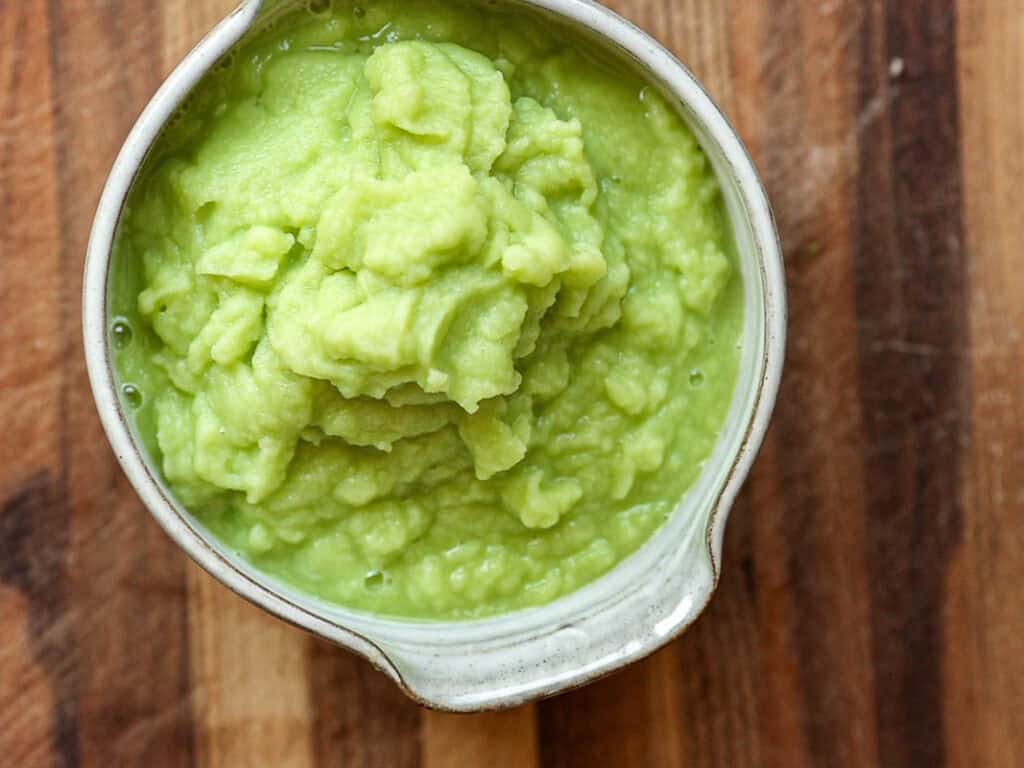 A small, white ceramic bowl filled with creamy broccoli puree on a wooden surface. The avocado has a smooth, green texture and looks fresh and appetizing.